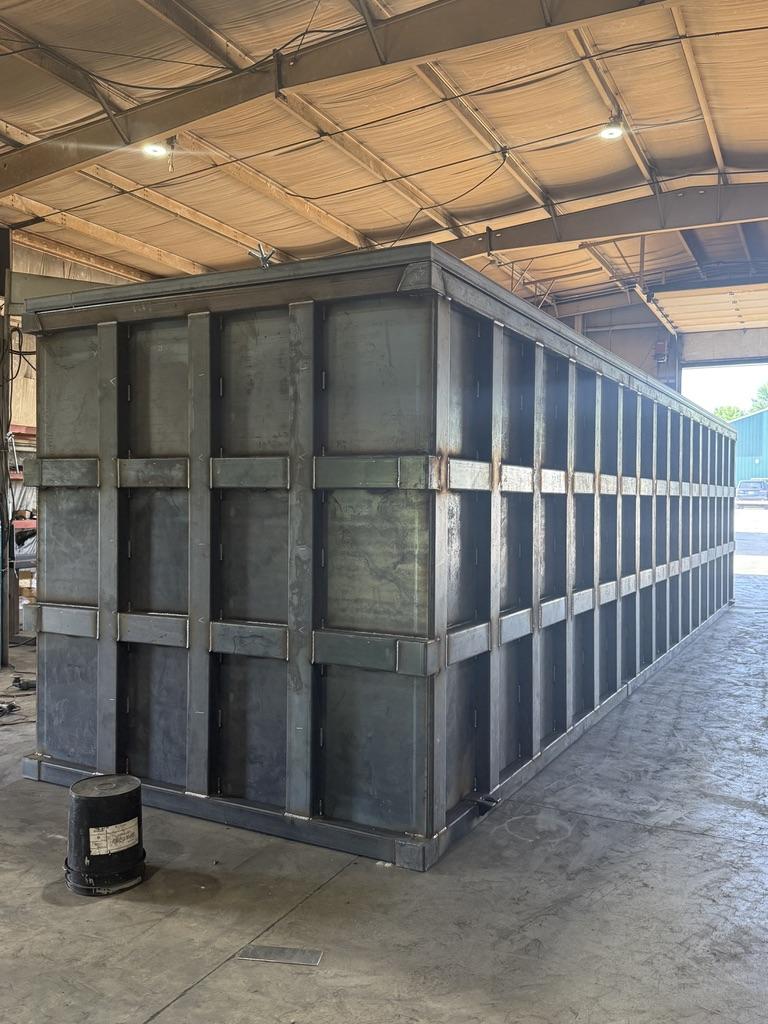 Angled view of a bare carbon-steel EnviroBox tank inside the shop showing the full rib grid